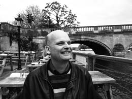 Jan  Hellbusch  in einem Biergarten an einem Fluss. Er lacht. Im Hintergrund erkennt man die massiven Bögen einer mit alten Bäumen bewachsenen Brücke.